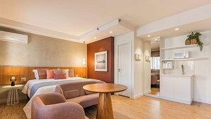 Una habitación de hotel moderna con una cama, una mesa redonda de madera, un área de descanso, una kitchenette y un baño visible a través de una puerta abierta.