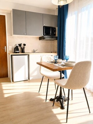 A compact kitchen with grey cabinets, a microwave, and a small fridge. There is a dining table with chairs and a blue curtain. - Le BlueAna – Studio moderne & confortable (Poitiers)