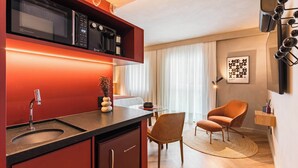 Une cuisine compacte équipée d’un four à micro-ondes, d’une machine à café et d’un évier. Un coin repas est aménagé avec une chaise et une petite table.