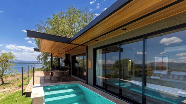 Un área de piscina exterior moderna con vista al mar, terraza de madera y zona de estar con paredes de cristal.