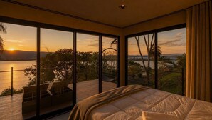 Un dormitorio con una cama grande, vistas a una puesta de sol sobre el agua y un balcón con mobiliario exterior.
