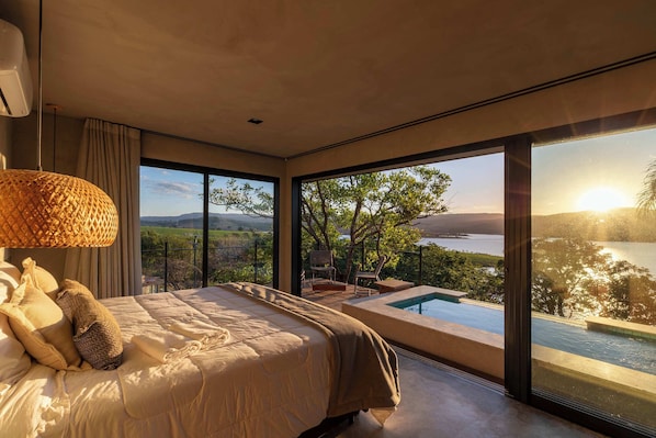 A modern bedroom with a large bed, a view of a lake, and an infinity pool.