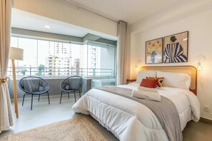 A hotel room with a large bed, two chairs, a balcony with a view of buildings, and artwork on the wall. - Studio Completo a 5 min do Nubank Parque | MP1410 (Perdizes)