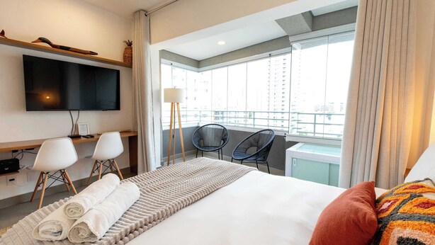 Ein Hotelzimmer mit Bett, Fernseher, Schreibtisch mit Stühlen und einem Balkon mit Stadtblick.