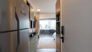 Eine schmale Küche mit einem Edelstahlkühlschrank, einem kleinen Essbereich mit zwei Stühlen und Blick auf Gebäude durch ein Fenster.