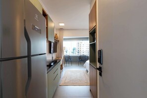 A narrow kitchen with a stainless steel refrigerator, a small dining area with two chairs, and a view of buildings through a window. - Studio Completo a 5 min do Nubank Parque | MP1410 (Perdizes)
