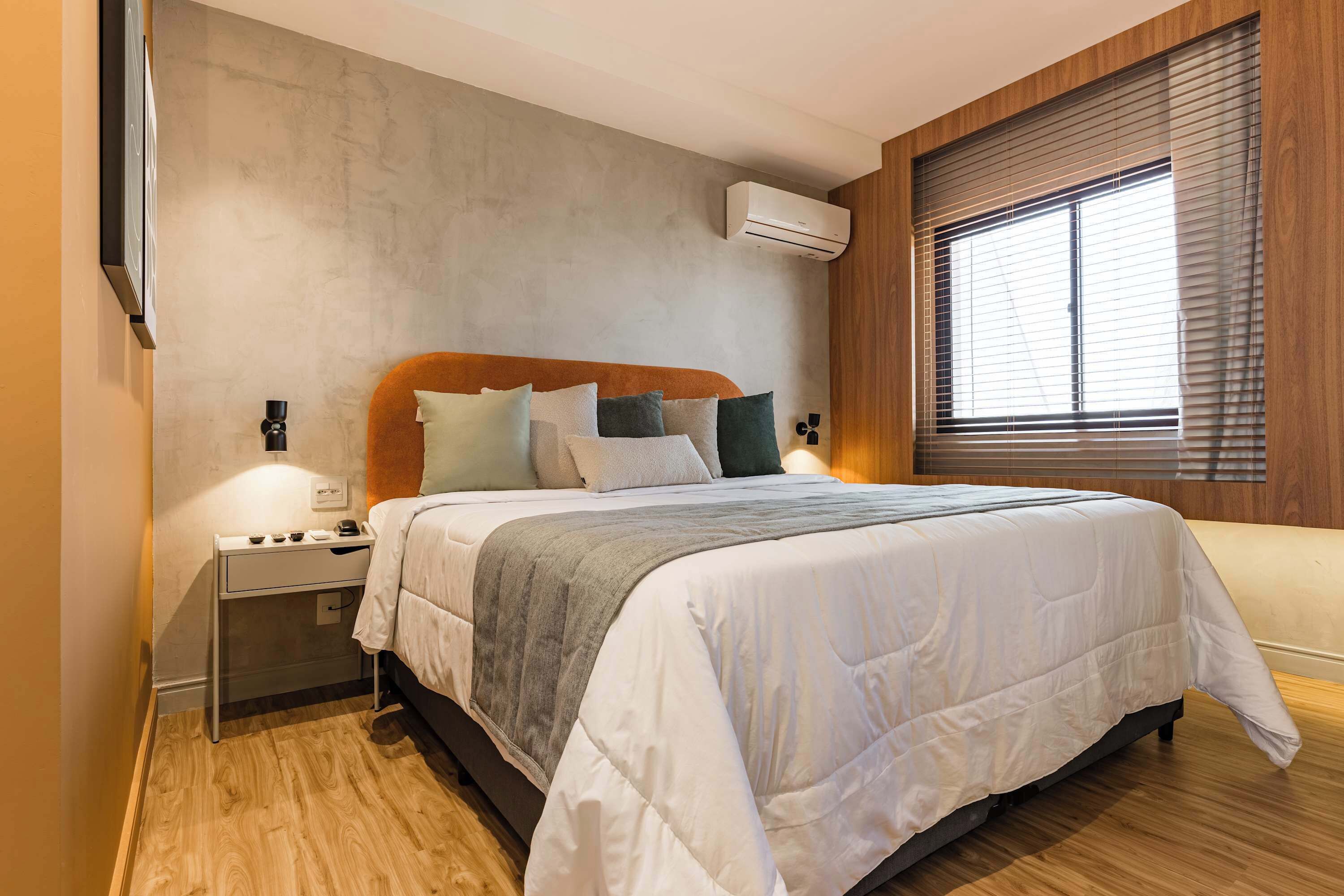 A bedroom with a bed, bedside tables, a wall-mounted air conditioner, and a window with blinds.