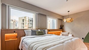 Une chambre moderne avec un grand lit, deux tables de chevet, une tête de lit en bois et une vue sur les immeubles à travers de grandes fenêtres.