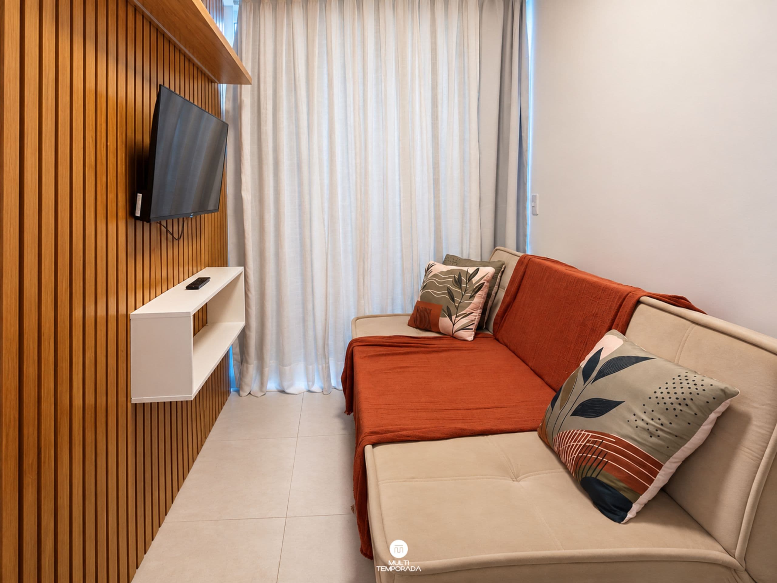Una habitación moderna con un panel de madera en la pared, un televisor montado, un sofá con cojines y una ventana con cortinas.