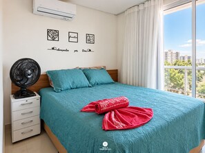 Et soveværelse med en seng, en ventilator, et natbord, en kommode og et vindue med udsigt til bygninger og træer.