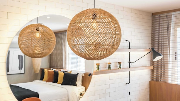 Una camera d'albergo moderna con uno specchio rotondo, un letto con molti cuscini, una lampada in vimini sferica, una scrivania con una lampada e una finestra con persiane.