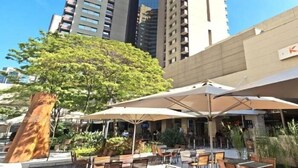 Un edificio alto con una sección cilíndrica, rodeado de mesas y sillas al aire libre con sombrillas.
