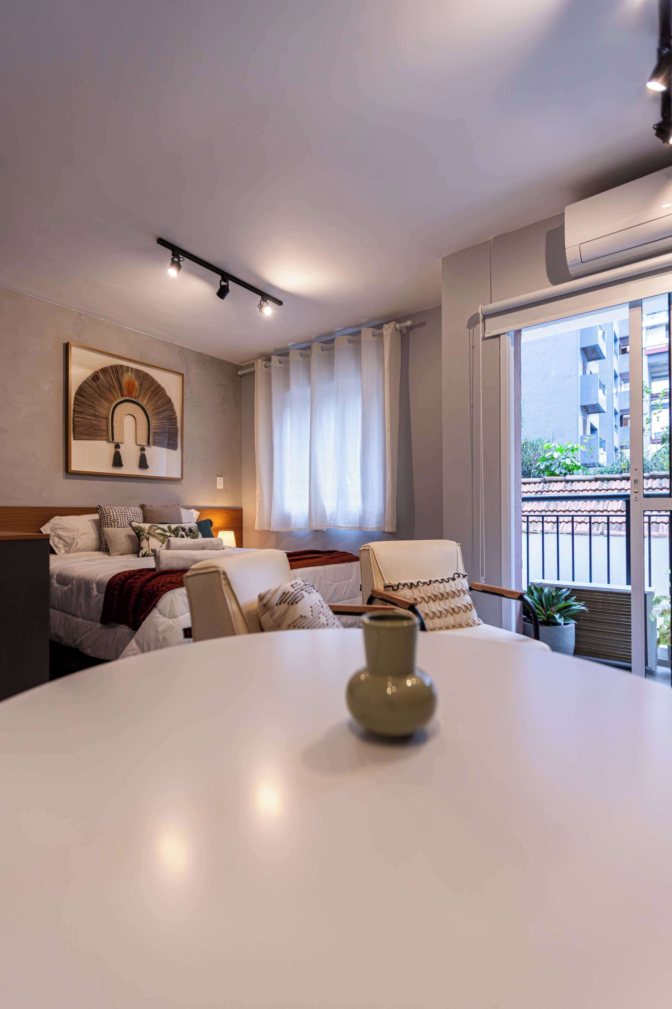 A hotel room with a bed, bedside table, lamp, potted plant, and a view of the outside.
