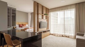 A modern hotel room with a large flat-screen TV, a wooden shelving unit, a bed with white bedding, a seating area with a chair, and a large window with sheer curtains. - #VN1609 | Cyclinn Vila Olímpia (Vila Olímpia)