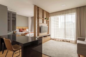 Ein modernes Hotelzimmer mit einem großen Flachbildfernseher, einer Holzeinrichtung, einem Bett mit weißer Bettwäsche, einer Sitzecke mit einem Sessel und einem großen Fenster mit durchsichtigen Vorhängen.