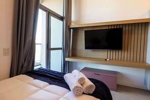 A hotel room with a bed, a flat-screen TV mounted on a wooden panel, a small pink cabinet, and a window with curtains. - Studio Completo a 100m do Metrô Brooklin | HM1113 (Brooklin)