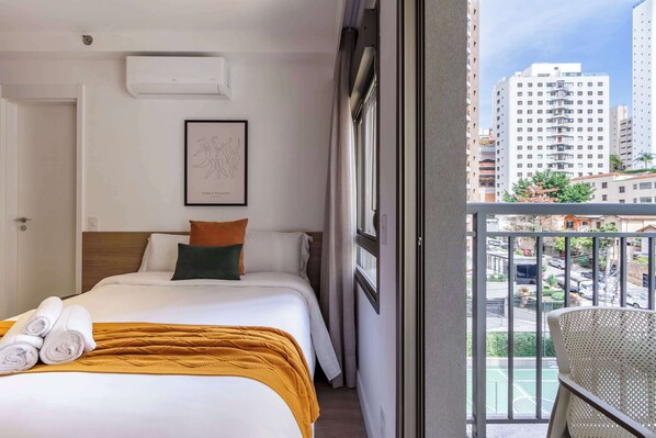 Une chambre d’hôtel avec un lit, un tableau encadré, une fenêtre avec des rideaux et un balcon avec une chaise.
