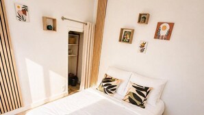 Ein ordentlich bezogenes Bett mit weißen Leinen und einem zusammengerollten Handtuch, ein Holzboden, eine gewebte Matte und ein kleiner Holzschemel.
