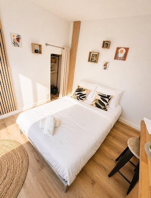 A neatly made bed with white linens and a rolled towel, a wooden floor, a woven mat, and a small wooden stool. - Le Petit Jean Alexandre – Studio centre-ville (Poitiers)