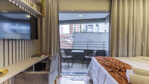 Hotellihuone, jossa on sänky, työpöytä, televisio, tuoleilla varustettu parveke ja näkymä rakennuksiin.