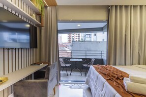 A hotel room with a bed, a desk, a TV, a balcony with chairs, and a view of buildings. - Studio Completo a 5 min do Nubank Parque | MP302 (Perdizes)