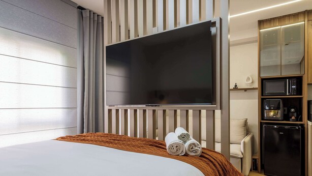 Una camera d'hotel con un letto, una TV a muro, un angolo cottura con microonde e frigorifero, e una finestra con tende.