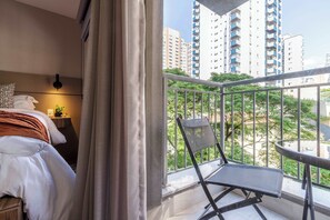 A hotel room with a bed, a nightstand, a lamp, a window with a view of high-rise buildings and greenery. - Studio Completo a 5min do Allianz Parque | GP33 (Perdizes)