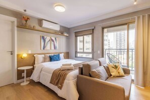 Una habitación de hotel moderna con cama, sofá, mesita de noche, obras de arte y un balcón con vistas.