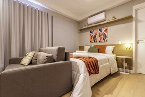 A hotel room with a sofa, a bed with white bedding, a small table with a lamp, and an air conditioning unit on the wall. - Ape Pet Friendly a 5 min do Nubank Parque GP55 (Perdizes)