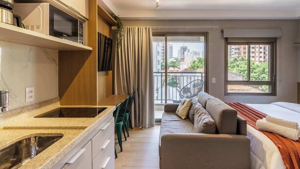 A modern hotel room with a kitchen area, a balcony with a view of the city, and a cozy seating area. - Ape Pet Friendly a 5 min do Nubank Parque GP55 (Perdizes)