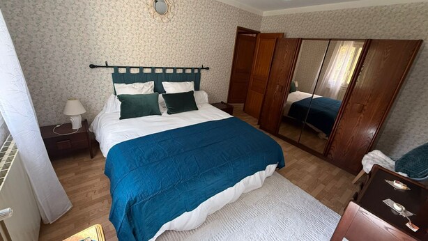 A bedroom with a bed, bedside table, lamp, mirror, wardrobe, and a small table with items on it. - The House of Seasons (Apt)