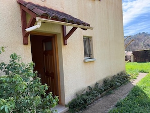 A building with a wooden door, a window with a metal frame, and a small garden area with greenery and a gravel path. - The House of Seasons (Apt)