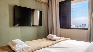 A hotel room with a bed, a TV mounted on the wall, a window with a city view, and a towel on the bedside table. - Flat a 300m do Metrô Consolação | BC1505 (Bela Cintra)