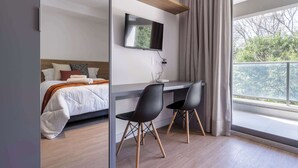 A hotel room with a bed, a desk with two chairs, a flat-screen TV, and a large window with curtains. - Studio Completo a 200m do Nubank Parque | AP3004 (Perdizes)