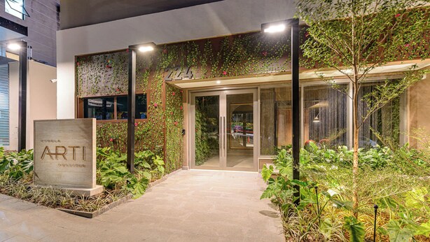 A modern building entrance with a sign reading 'ARTI,' surrounded by lush greenery and a paved pathway. - Studio Completo a 200m do Nubank Parque | AP3004 (Perdizes)