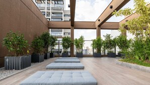 A rooftop terrace with seating areas, potted plants, and a view of a modern building. - Studio Completo a 100m do Metrô Brooklin | HM1108 (Brooklin)