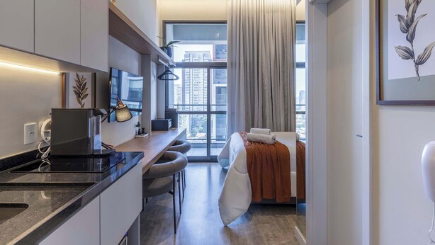 A modern hotel room with a kitchen area, a large window with a view, and a bed with a white pillow and orange blanket. - Studio Completo a 100m do Metrô Brooklin | HM1108 (Brooklin)