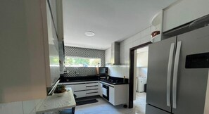 A modern kitchen with white cabinets, a stainless steel refrigerator, a microwave, and a window with a view of greenery. - Recanto Jacuípe Prime c Piscina Camaçari (Camaçari)