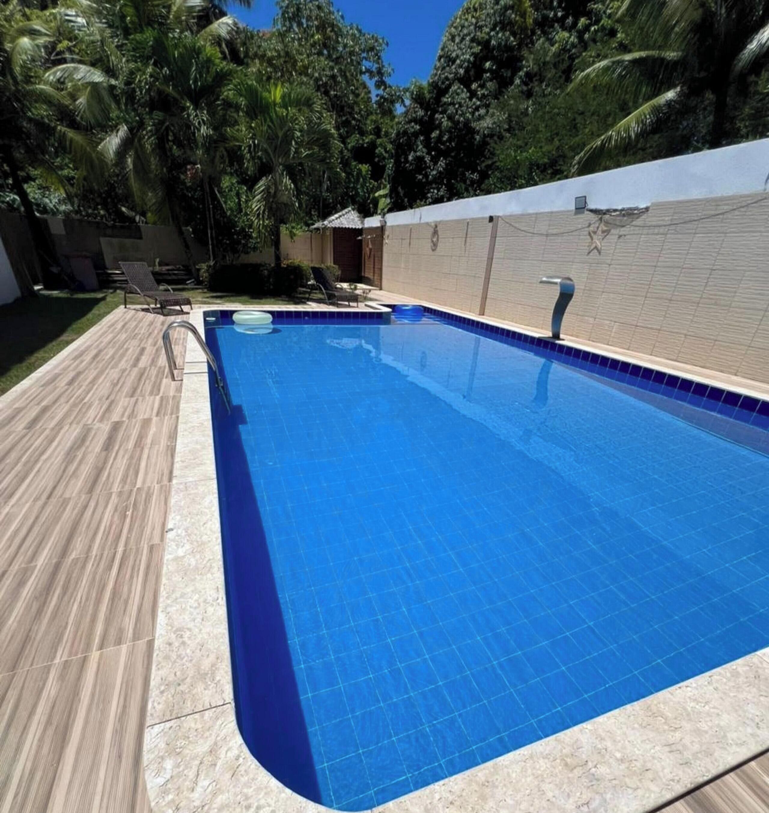 A rectangular swimming pool with a tiled edge, surrounded by a wooden deck and a white wall with decorative elements.