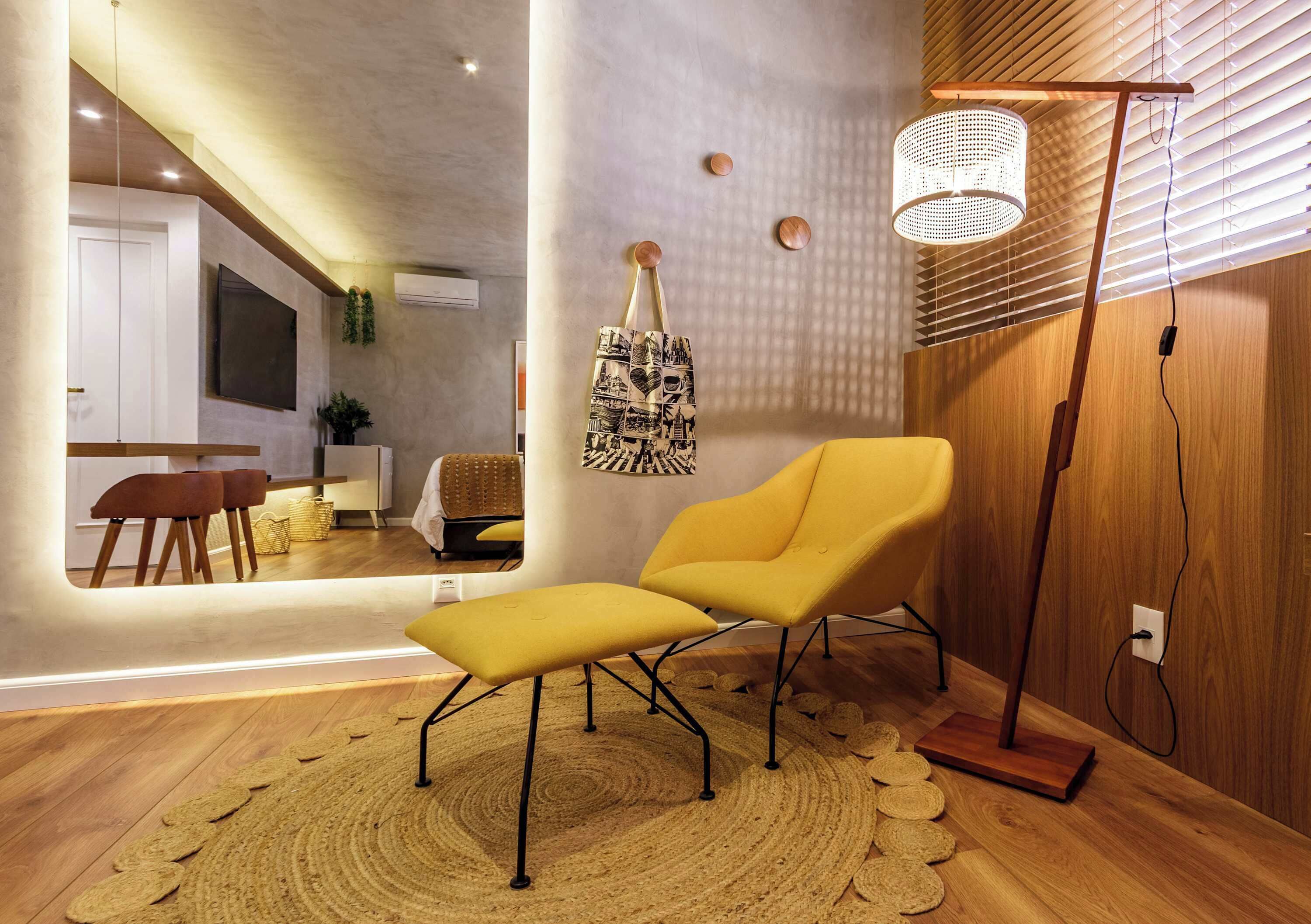 Een moderne zithoek met een gele fauteuil, een geweven vloerkleed, een vloerlamp en een grote spiegel die de kamer weerspiegelt.