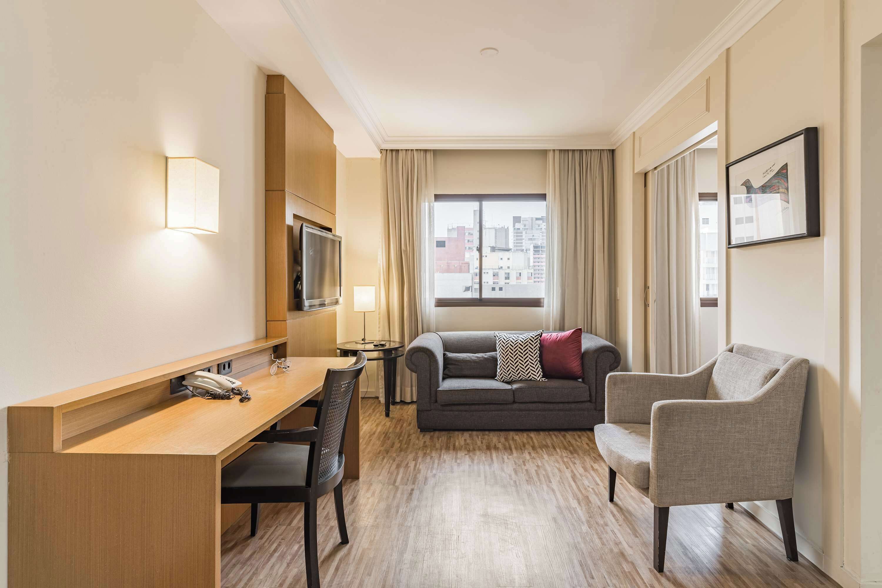 Ein Hotelzimmer mit einem Holztisch, einem Sofa, einem Sessel, einem Fernseher und einem Fenster mit Vorhängen.