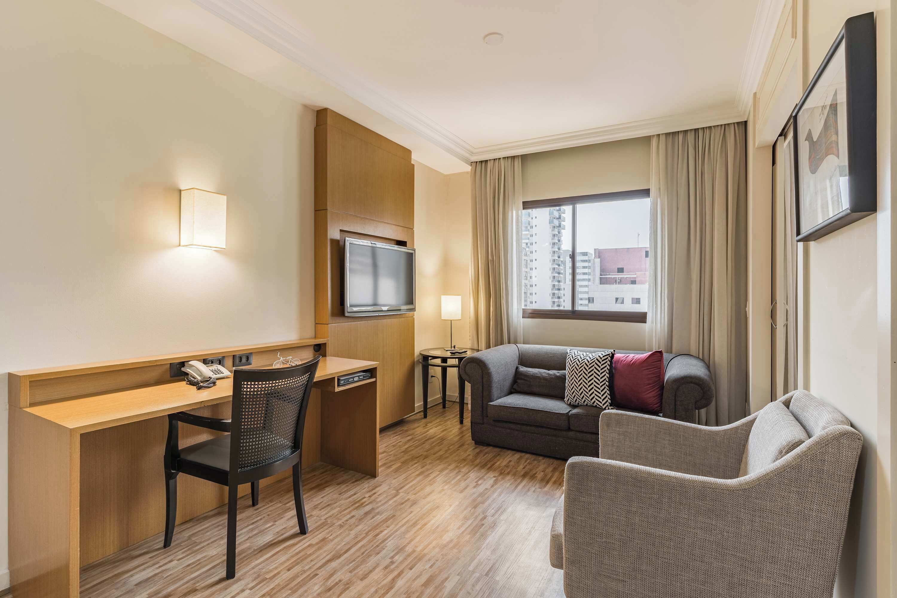 Ein Hotelzimmer mit Schreibtisch, Fernseher, einer Couch und einem fenster mit Vorhängen.