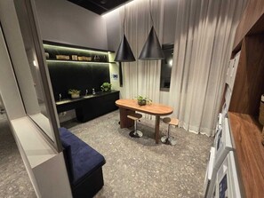 A modern room with a wooden dining table, two bar stools, a blue bench, a black pendant light, a wall-mounted shelf with plants, and a large window with curtains. - Studio Completo 500m do Metrô Borba Gato | #AD201 (Brooklin)