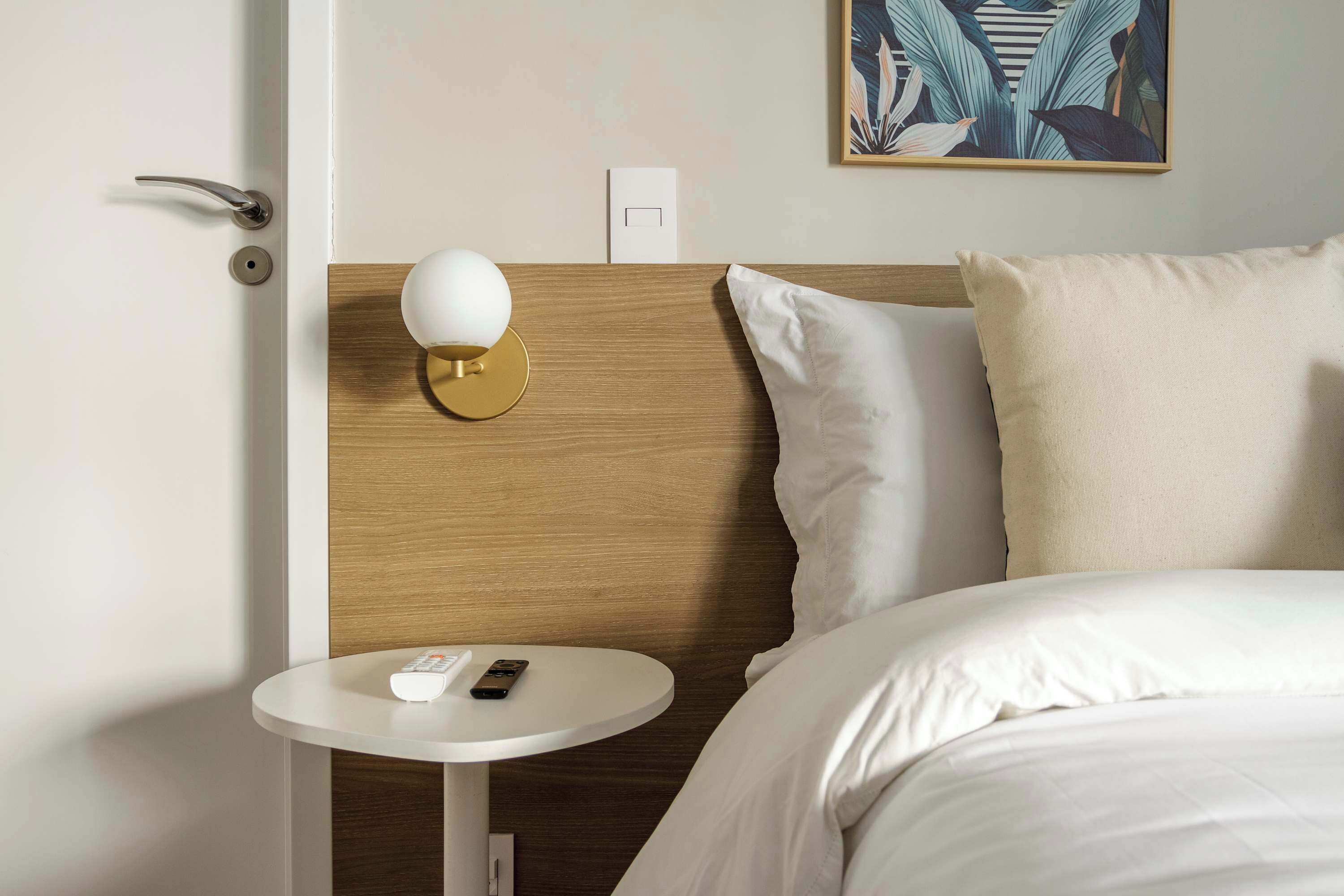 Ein Hotelzimmer mit Bett, Nachttisch mit Fernbedienung, Wandlampe und Steckdose.