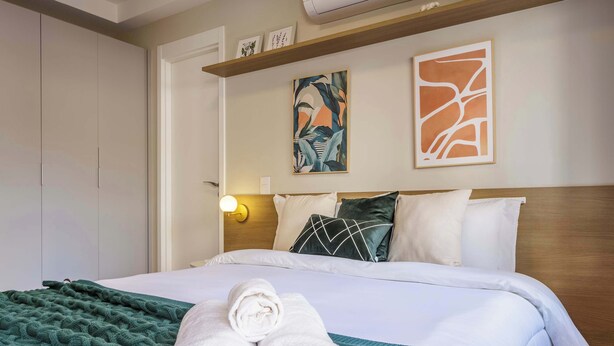 Habitación de hotel con cama, manta verde, dos toallas enrolladas, cabecero de madera y dos cuadros colgados en la pared.