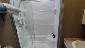 Eine Dusche mit Glastür, eine weiße Fliesenwand und eine kleine Ablage für Pflegeprodukte.