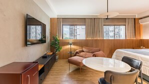 Kompakti hotellihuone, jossa on sänky, pieni pöytä, sohva, televisio, kasvi ja työpöytä tuolilla.