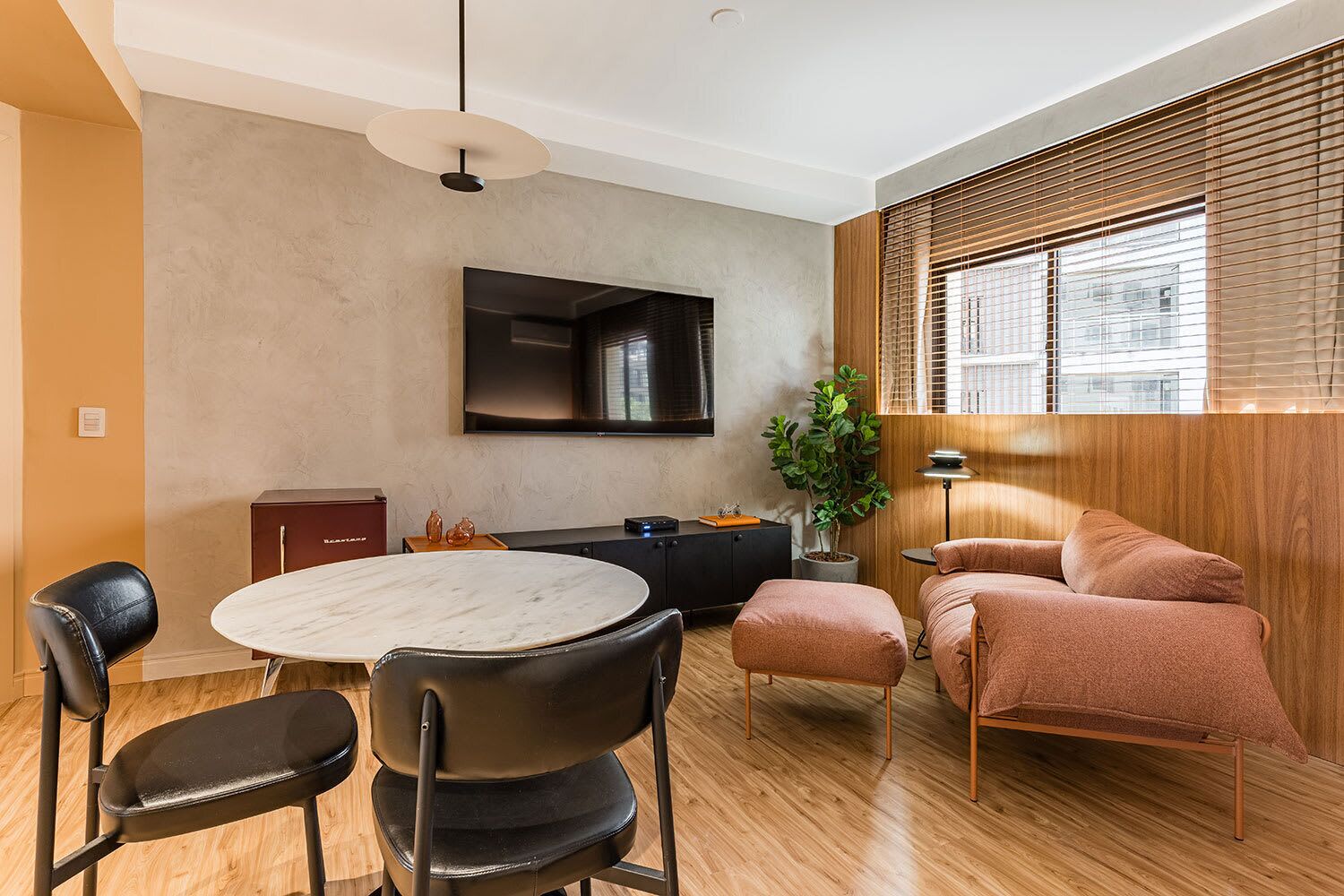 A modern living room with a round dining table, black chairs, a sofa, a TV mounted on the wall, and a potted plant.