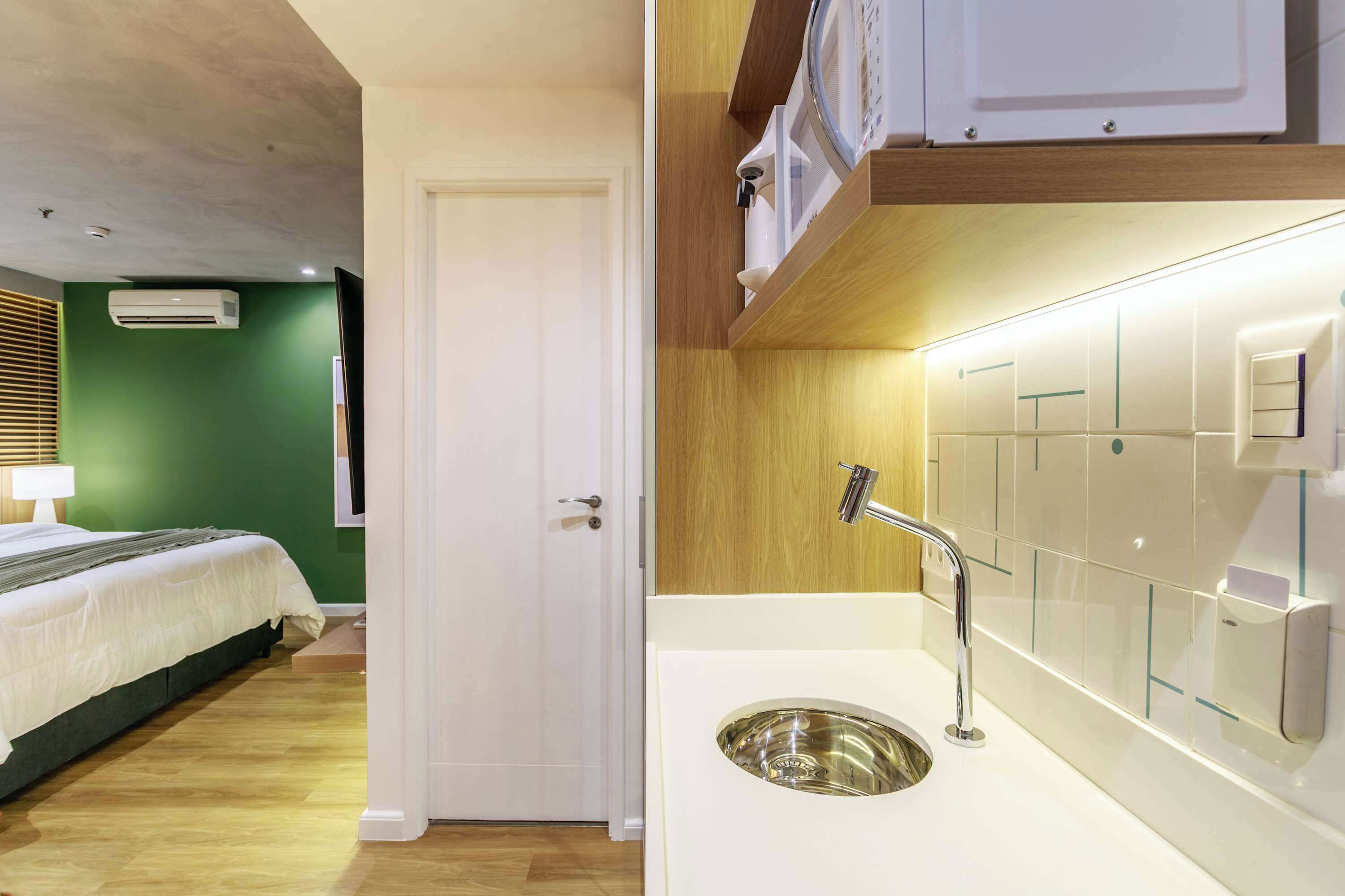 A hotel room with a bed, a white door, a sink with a mirror, and a wall-mounted air conditioner.