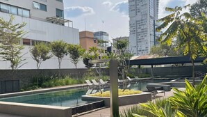 Un área de piscina en la azotea con una piscina rectangular, sillas de descanso y un edificio alto al fondo.